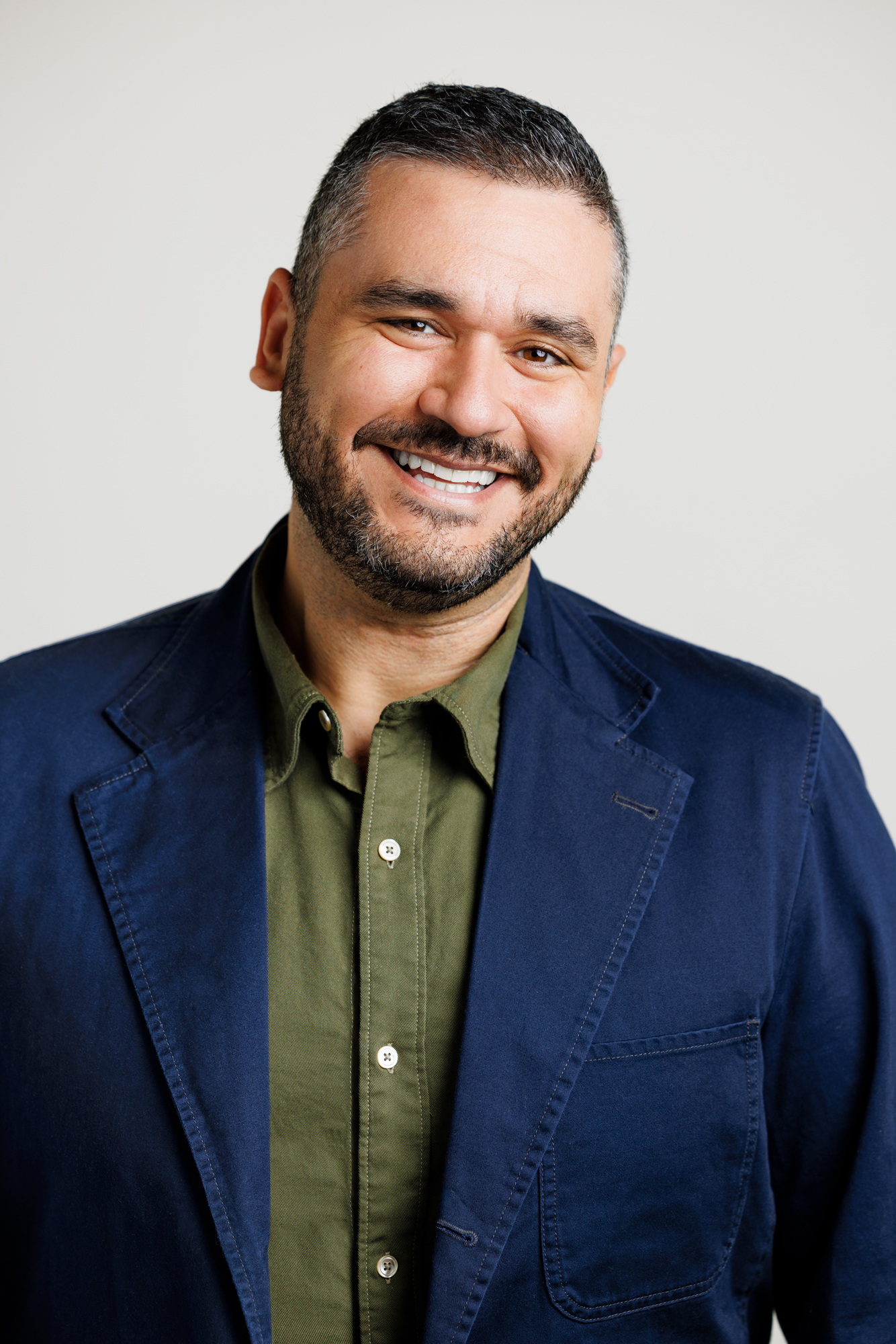 Portrait of Donesh Olyaie in a navy jacket and olive shirt against a pale grey ground
